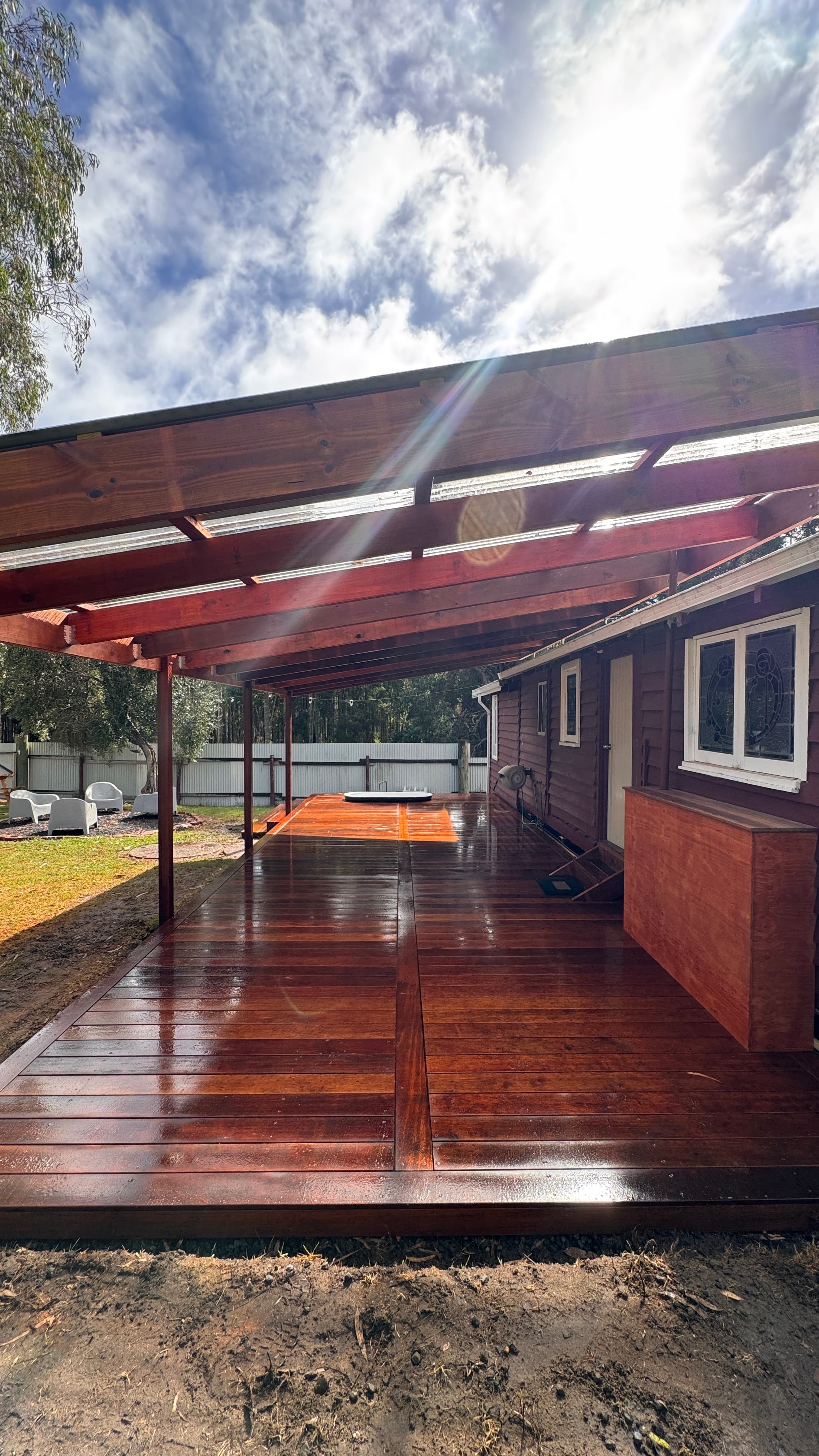Polished reddish-brown wooden deck under a pergola, reflecting bright sunlight next to a house.