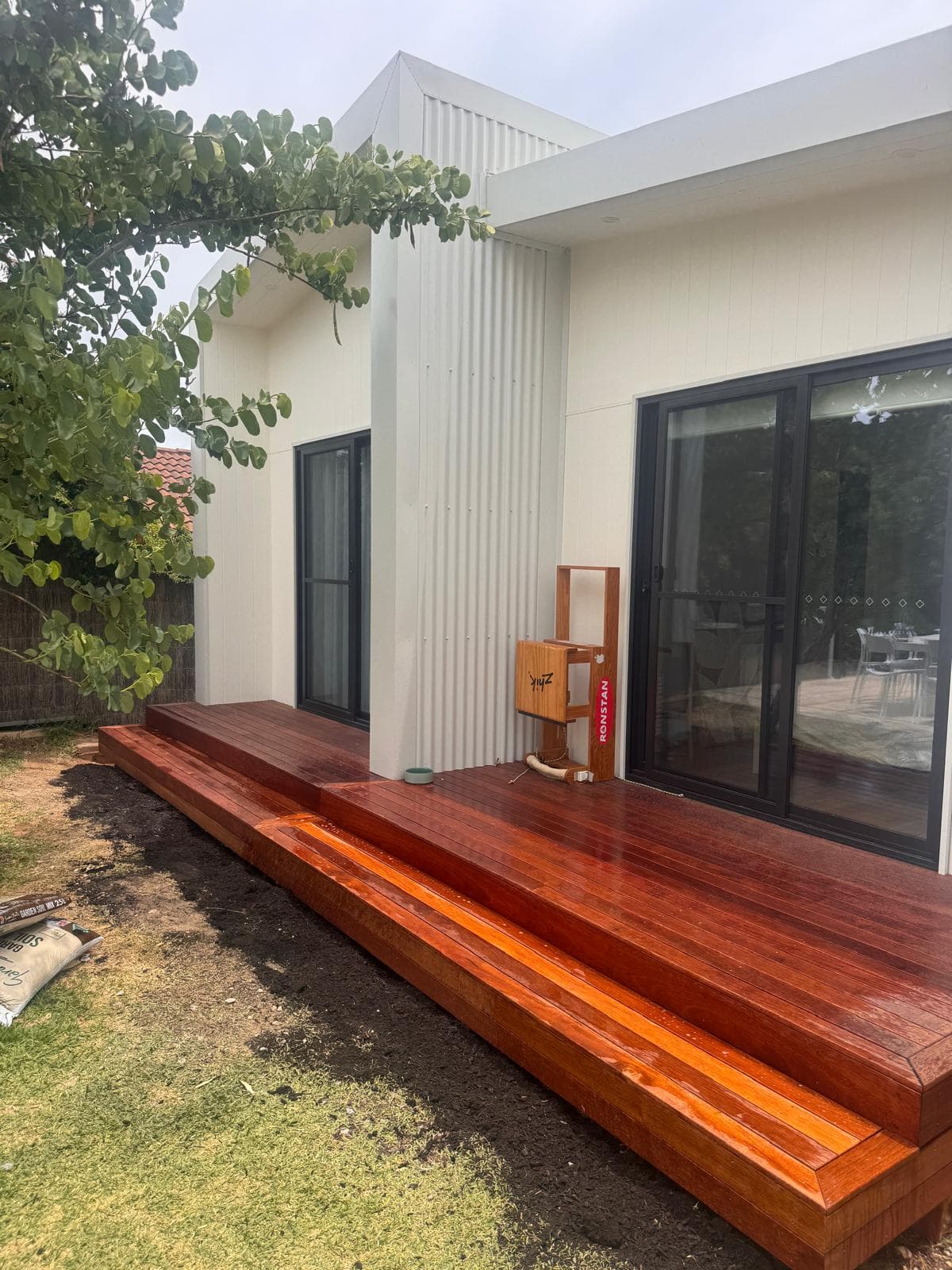 Polished reddish-brown wooden deck with steps outside a modern white house with black sliding doors.