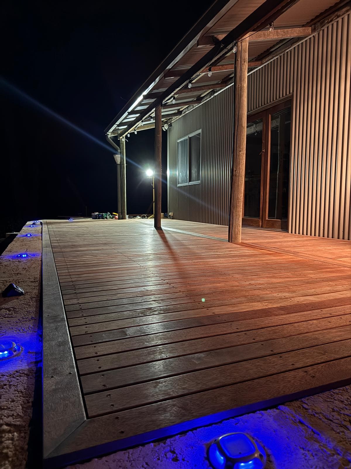Wooden deck at night with blue LED ground lights next to a corrugated metal building.