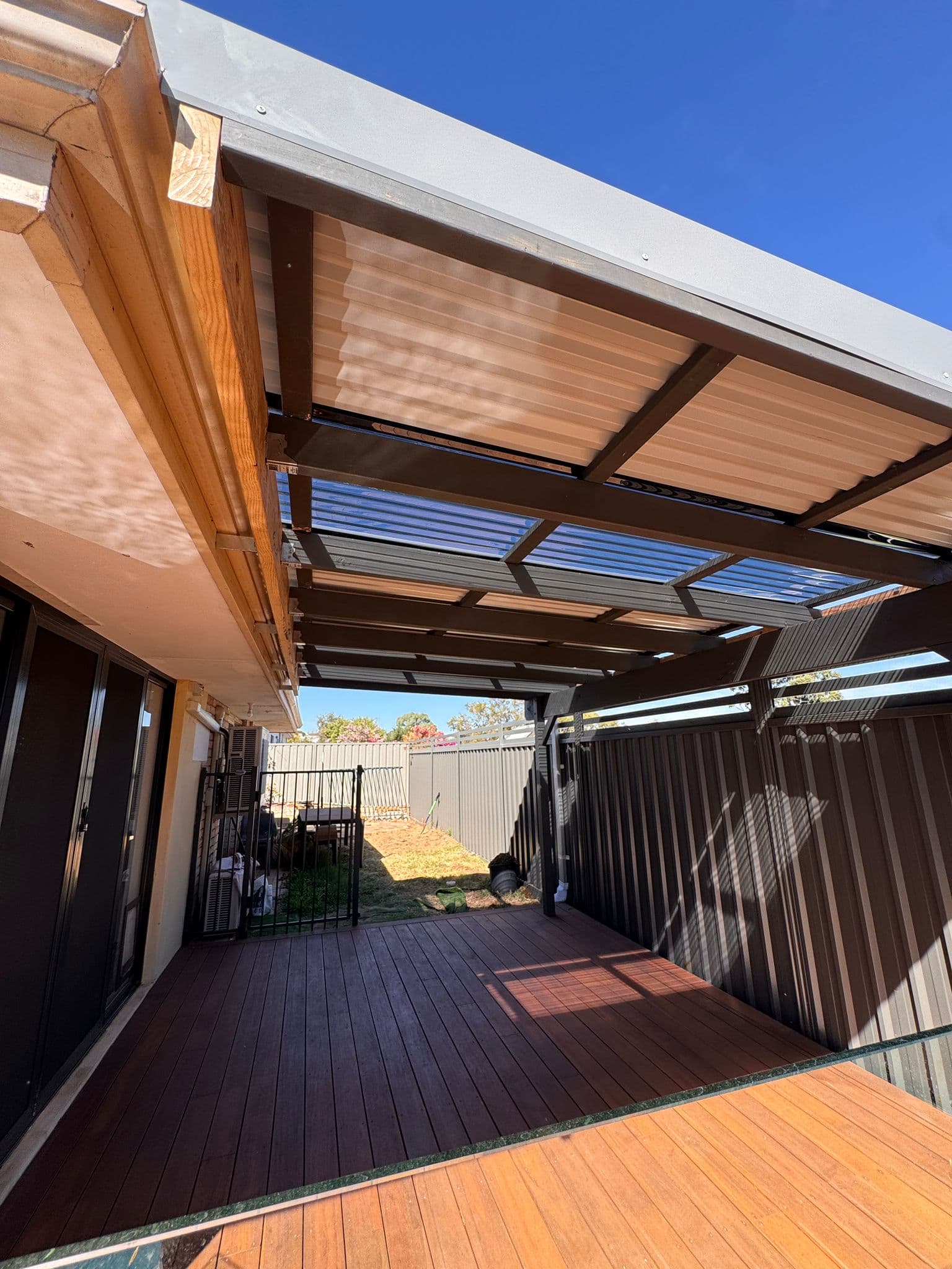 Dark wooden deck under a metal patio cover with translucent panels in a sunny backyard.