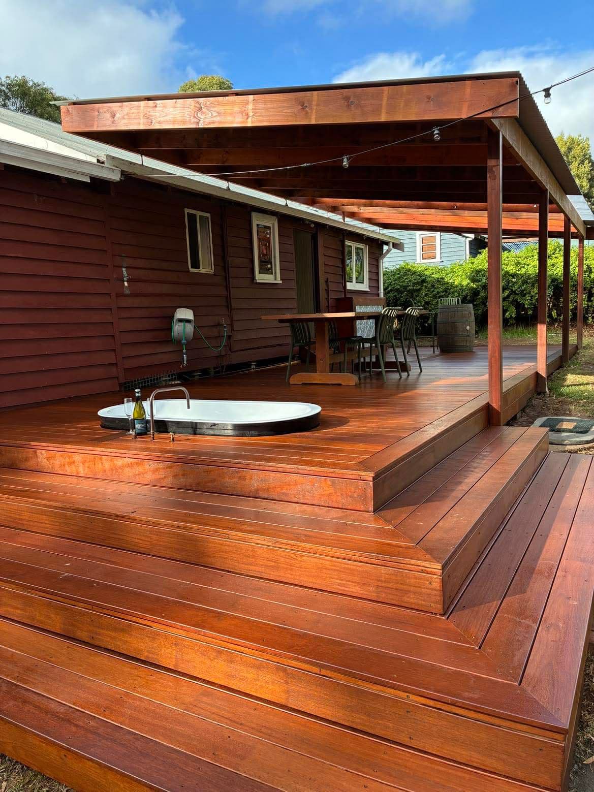 Reddish-brown tiered wooden deck featuring a sunken white bathtub, wine bottle, and covered pergola.