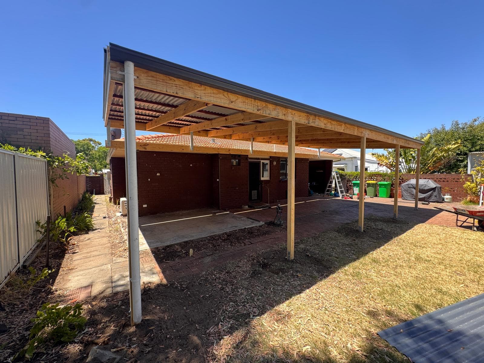 Wooden patio structure with metal roofing attached to a brick house in a sunny backyard.