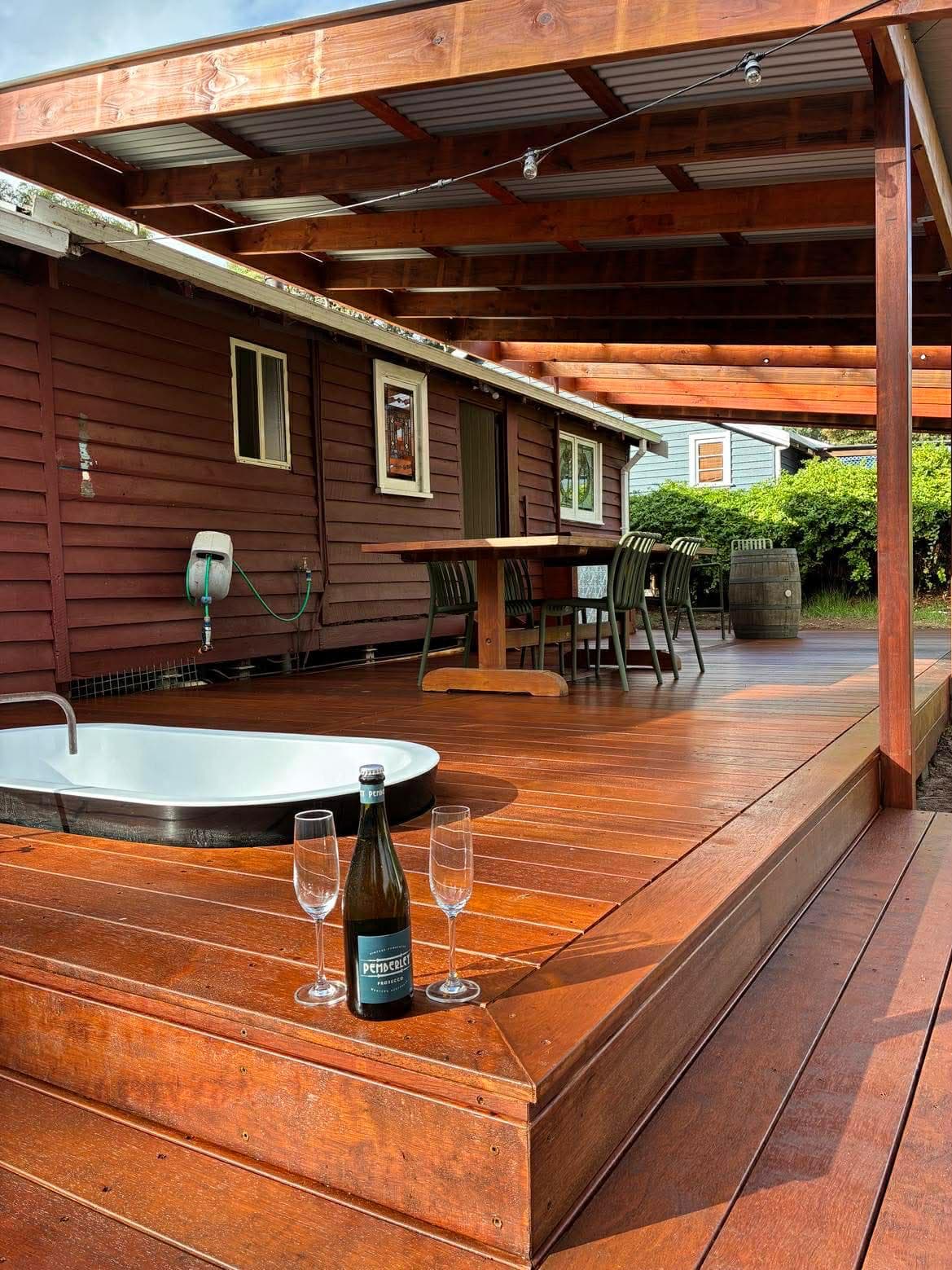 Wooden deck with a built-in bathtub, prosecco, and dining area under a covered pergola.
