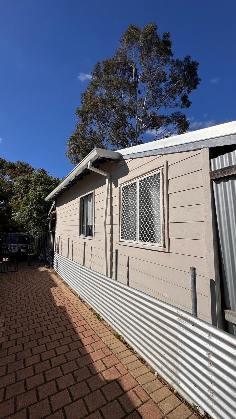 Beige house with horizontal siding and corrugated metal fencing along a brick-paved walkway.